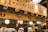 A cozy restaurant interior with a warm, ambient lighting and a wooden panel adorned with masks and Japanese calligraphy. Lanterns hang from the ceiling, and a chef is visible behind a counter, interacting with customers.