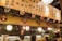 A cozy restaurant interior with a warm, ambient lighting and a wooden panel adorned with masks and Japanese calligraphy. Lanterns hang from the ceiling, and a chef is visible behind a counter, interacting with customers.