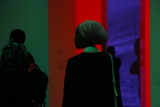 A silhouette of a person wearing a headscarf stands in front of a colorful illuminated installation. The background features a blend of red and blue hues, creating a striking contrast. Other figures are faintly visible, partially obscured by shadow.