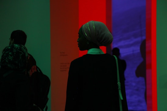 A silhouette of a person wearing a headscarf stands in front of a colorful illuminated installation. The background features a blend of red and blue hues, creating a striking contrast. Other figures are faintly visible, partially obscured by shadow.