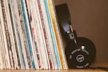 A collection of vinyl records stands upright, showcasing various album covers with diverse text and colors. Beside them, a pair of black headphones with visible branding rests against the records.