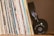 A collection of vinyl records stands upright, showcasing various album covers with diverse text and colors. Beside them, a pair of black headphones with visible branding rests against the records.