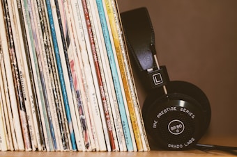 A collection of vinyl records stands upright, showcasing various album covers with diverse text and colors. Beside them, a pair of black headphones with visible branding rests against the records.