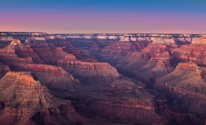 The image captures a breathtaking view of the Grand Canyon, showcasing its layered, multi-colored rock formations and vast, rugged landscape. The sky above transitions from a pale, serene pink near the horizon to a deeper blue above, enhancing the natural beauty of the canyon.