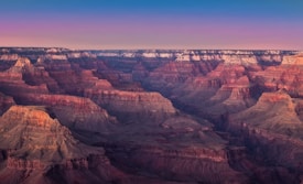 The image captures a breathtaking view of the Grand Canyon, showcasing its layered, multi-colored rock formations and vast, rugged landscape. The sky above transitions from a pale, serene pink near the horizon to a deeper blue above, enhancing the natural beauty of the canyon.