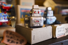 A vintage-style robot toy is placed inside a cardboard display box. The robot has a silver metallic finish, red eyes, and a retro design. In the background, another similar robot is visible along with colorful toys and signage with Japanese text.