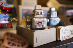 A vintage-style robot toy is placed inside a cardboard display box. The robot has a silver metallic finish, red eyes, and a retro design. In the background, another similar robot is visible along with colorful toys and signage with Japanese text.