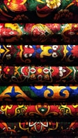 Detailed, vibrant fabric with intricate and colorful patterns. The fabric features an array of motifs, including floral designs and geometric shapes, arranged in horizontal rows. The varied textures and rich colors create a visually striking and dynamic composition.
