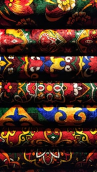 Detailed, vibrant fabric with intricate and colorful patterns. The fabric features an array of motifs, including floral designs and geometric shapes, arranged in horizontal rows. The varied textures and rich colors create a visually striking and dynamic composition.