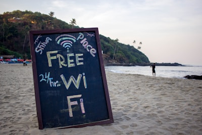 A chalkboard sign is displayed prominently on a sandy beach, advertising 'Free Wi-Fi 24 hr.' with colorful text. In the background, there are green hills with palm trees, and a person is standing by the shoreline looking out at the ocean. The scene is tranquil with a clear sky.