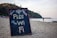 A chalkboard sign is displayed prominently on a sandy beach, advertising 'Free Wi-Fi 24 hr.' with colorful text. In the background, there are green hills with palm trees, and a person is standing by the shoreline looking out at the ocean. The scene is tranquil with a clear sky.
