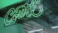 A neon sign with the word 'Court' in vibrant green light is mounted on an industrial ceiling. The background includes exposed metal beams and a partially visible suspended lamp, creating an urban and modern atmosphere.
