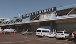 A large industrial-looking building with a substantial sign reading 'Sydney Fish Market' prominently displayed at the top. Several loading docks are visible, labeled with numbers, and there are multiple vehicles, including white vans and a car, parked in front. People are walking near the parked vehicles. The weather appears clear and the overall atmosphere is busy yet organized.