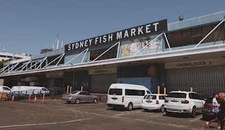 A large industrial-looking building with a substantial sign reading 'Sydney Fish Market' prominently displayed at the top. Several loading docks are visible, labeled with numbers, and there are multiple vehicles, including white vans and a car, parked in front. People are walking near the parked vehicles. The weather appears clear and the overall atmosphere is busy yet organized.