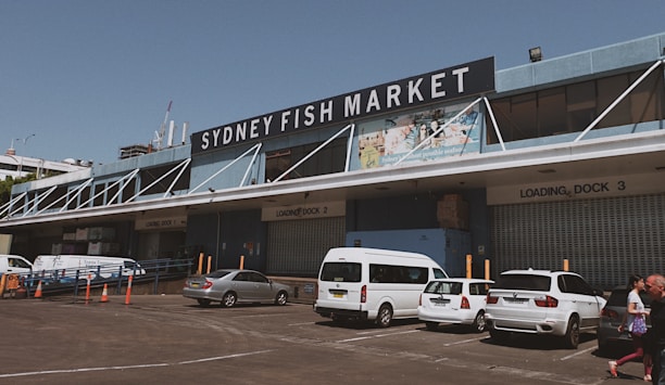 A large industrial-looking building with a substantial sign reading 'Sydney Fish Market' prominently displayed at the top. Several loading docks are visible, labeled with numbers, and there are multiple vehicles, including white vans and a car, parked in front. People are walking near the parked vehicles. The weather appears clear and the overall atmosphere is busy yet organized.