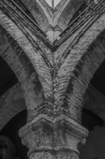 An upward view of a stone or brick architectural structure featuring arches and detailed masonry work. The image is captured in black and white, emphasizing the texture and age of the construction.