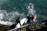 Surfers are entering the ocean, carrying their boards down a rocky staircase. The water is choppy with visible waves and a surfer is already paddling out. They are all wearing wetsuits, indicating cooler water temperatures.
