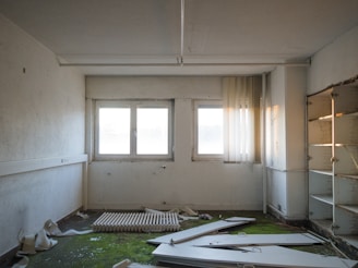 A neglected, empty room with peeling paint and a broken radiator lying on a green carpet stained with dirt. The windows are covered with dusty blinds, and there are broken doors and debris scattered across the floor. Shelves in the corner appear rusted and abandoned.