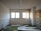 A neglected, empty room with peeling paint and a broken radiator lying on a green carpet stained with dirt. The windows are covered with dusty blinds, and there are broken doors and debris scattered across the floor. Shelves in the corner appear rusted and abandoned.