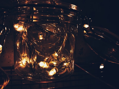 A glass jar filled with glowing string lights, emitting a warm yellowish glow. The wire and bulbs are intertwined, creating a tangled yet organized appearance inside the jar. Ambient lighting is dim, highlighting the brightness of the lights within the jar.