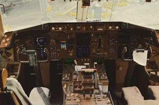A cockpit filled with numerous instruments, gauges, and controls. There are dual joysticks and a complex array of buttons and screens, set against a backdrop of some paper items clipped within reach. The view includes the exterior area of the airport visible through the cockpit windows.