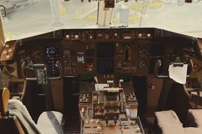 A cockpit filled with numerous instruments, gauges, and controls. There are dual joysticks and a complex array of buttons and screens, set against a backdrop of some paper items clipped within reach. The view includes the exterior area of the airport visible through the cockpit windows.