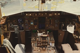 A cockpit filled with numerous instruments, gauges, and controls. There are dual joysticks and a complex array of buttons and screens, set against a backdrop of some paper items clipped within reach. The view includes the exterior area of the airport visible through the cockpit windows.