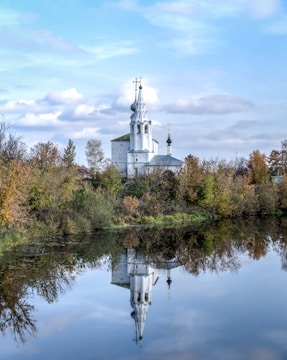 A white church with a tall spire and crosses is surrounded by autumn trees. The church is reflected in a calm body of water beneath it, creating a symmetrical image. The sky above is partly cloudy, adding a serene backdrop to the scene.