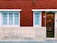 A red brick building facade featuring two windows with white frames and blue security bars on the left, and a brown wooden door with a mailbox slot on the right. The wall also has a small decorative plant above the door number sign.