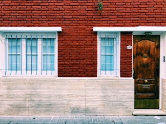 A red brick building facade featuring two windows with white frames and blue security bars on the left, and a brown wooden door with a mailbox slot on the right. The wall also has a small decorative plant above the door number sign.