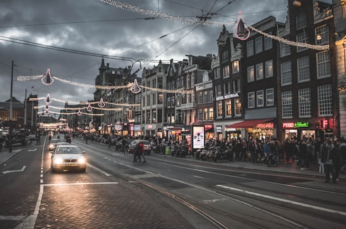 A bustling urban street scene at dusk with numerous people walking along the sidewalks and vehicles driving on the road. The street is adorned with festive string lights. The architecture features a row of multi-story buildings with shops and restaurants, some displaying illuminated signs.