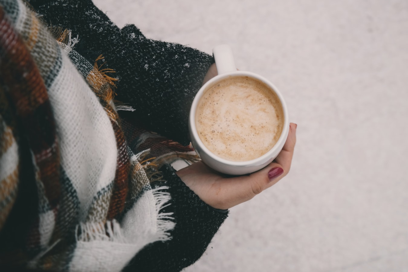 view from above of white woman holding a cup of cappuccino, she's dressed for winter and only her arms and hands are visible, with snowy ground as the background