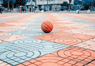 A basketball rests in the center of a colorful patterned courtyard. The surface is composed of red and blue tiles with intricate designs. In the background, a few people are visible near playground equipment and buildings.