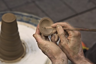 Hands skillfully sculpting a small clay object using a thin tool, with a partially shaped clay piece on a rotating pottery wheel nearby.