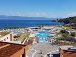 A picturesque coastal resort features multiple swimming pools surrounded by palm trees and sun loungers. The buildings have modern architecture, with terraces overlooking the vibrant blue sea. The landscape includes lush greenery and manicured gardens. The backdrop of a serene ocean and distant mountains enhances the tranquil setting.