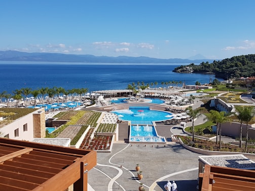 A picturesque coastal resort features multiple swimming pools surrounded by palm trees and sun loungers. The buildings have modern architecture, with terraces overlooking the vibrant blue sea. The landscape includes lush greenery and manicured gardens. The backdrop of a serene ocean and distant mountains enhances the tranquil setting.