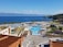 A picturesque coastal resort features multiple swimming pools surrounded by palm trees and sun loungers. The buildings have modern architecture, with terraces overlooking the vibrant blue sea. The landscape includes lush greenery and manicured gardens. The backdrop of a serene ocean and distant mountains enhances the tranquil setting.
