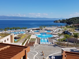 A picturesque coastal resort features multiple swimming pools surrounded by palm trees and sun loungers. The buildings have modern architecture, with terraces overlooking the vibrant blue sea. The landscape includes lush greenery and manicured gardens. The backdrop of a serene ocean and distant mountains enhances the tranquil setting.