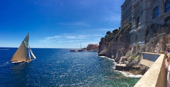 A classic sailboat glides across a sparkling blue sea near a rugged coastline with an ornate stone building perched atop the cliffs. The sky is a clear, bright blue, with a few wispy clouds. People are gathered along the edge of a walkway, gazing at the scenic view.