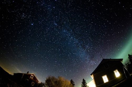 A night sky filled with countless stars stretches over a landscape featuring two houses. The Milky Way is visible, creating a captivating band of stars. One house is brightly lit from the inside, while the other remains in shadow. Sparse trees dot the horizon, and a subtle gradient of green suggests the presence of the aurora borealis.