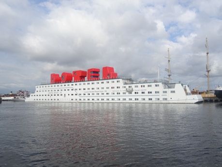 A large white ship is docked on a calm body of water. The structure has many windows and the word 'hotel' prominently displayed in big, red letters on the top. The ship is situated in a port with other masts and boats visible in the background, under a partly cloudy sky.