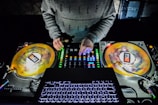 A DJ is working with vinyl turntables and a mixer in a dark setting. The DJ's hands are actively adjusting the controls on the mixer, and the spinning vinyl records are visible. The laptop keyboard is illuminated in the foreground.