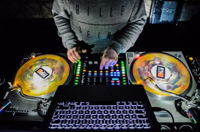 A DJ is working with vinyl turntables and a mixer in a dark setting. The DJ's hands are actively adjusting the controls on the mixer, and the spinning vinyl records are visible. The laptop keyboard is illuminated in the foreground.