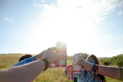 Friends laughing and clinking pop stream cans at a sunny outdoor picnic with vibrant decorations.