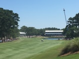 A golf course with a well-maintained green landscape is visible, surrounded by tall trees. A few people are gathered on the fairway, likely watching or participating in a golf event. In the background, grandstands are filled with spectators, and a crane with a camera is positioned for coverage. A pond is also visible, adding to the scenic beauty.