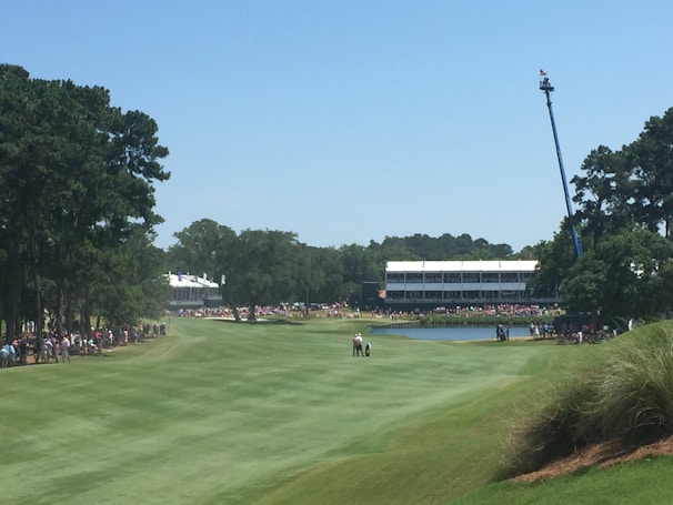A golf course with a well-maintained green landscape is visible, surrounded by tall trees. A few people are gathered on the fairway, likely watching or participating in a golf event. In the background, grandstands are filled with spectators, and a crane with a camera is positioned for coverage. A pond is also visible, adding to the scenic beauty.