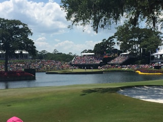 A large golf course with spectators gathered around, watching a game. Trees surround the area, which includes a water hazard and sand trap. The crowd is seated on grassy banks, under a clear sky with some clouds.
