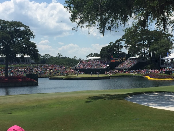 A large golf course with spectators gathered around, watching a game. Trees surround the area, which includes a water hazard and sand trap. The crowd is seated on grassy banks, under a clear sky with some clouds.