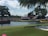 A large golf course with spectators gathered around, watching a game. Trees surround the area, which includes a water hazard and sand trap. The crowd is seated on grassy banks, under a clear sky with some clouds.