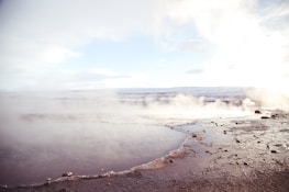 A geothermal landscape with a pool of water in the foreground surrounded by rock and soil. Steam rises gently from the surface, blending with the cloudy sky. The horizon shows distant mountains under a pale blue sky, creating a serene and expansive view.